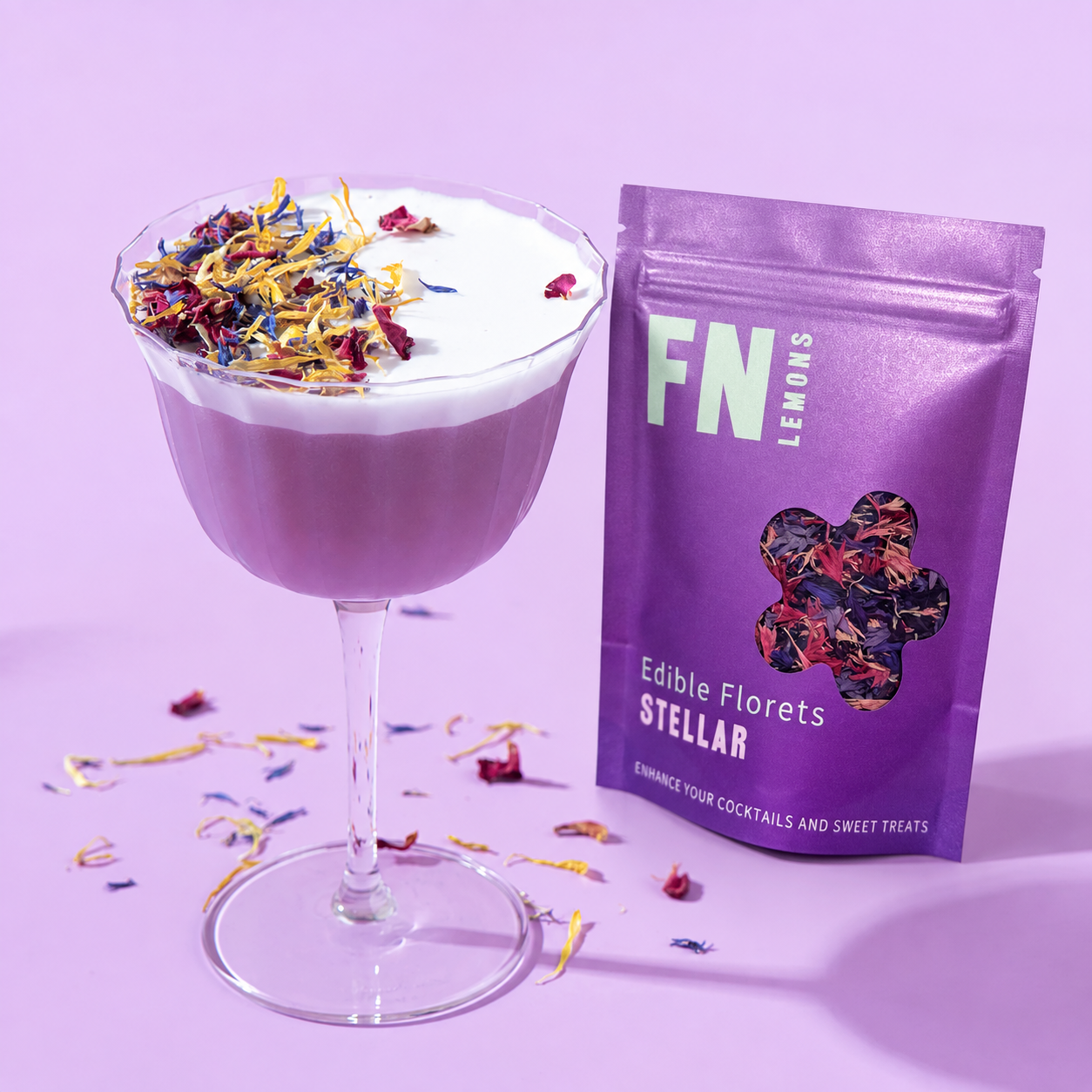 Load image into Gallery viewer, A purple cocktail in a coupe glass topped with white foam and edible flower petals, shown next to a purple stand-up pouch labeled 'FN Lemons Stellar Edible Florets'. The scene is styled on a light purple background with scattered flower petals.