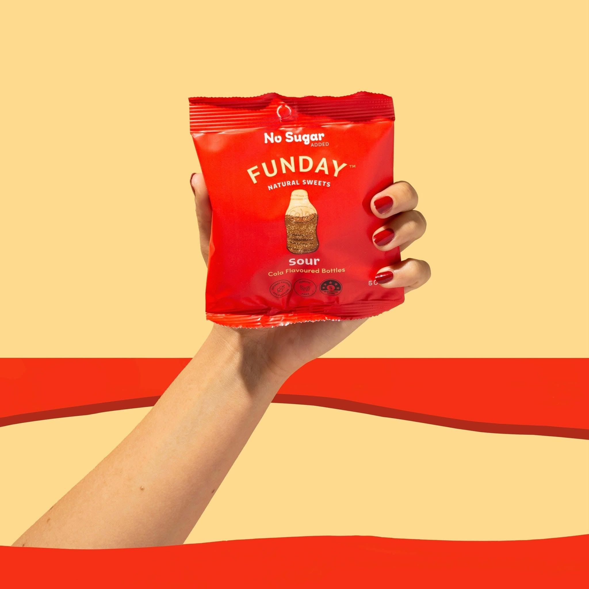 Load image into Gallery viewer, A hand with red nail polish holds a red bag of No Sugar Funday Cola Flavoured bottles Natural Sweets against a warm beige background with a bold red horizontal stripe.