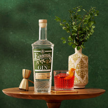 Load image into Gallery viewer, Bottle of Farmers Gin on a wooden table with a glass of red cocktail and a vase with greenery.