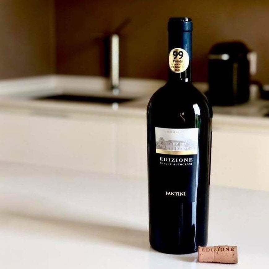 Load image into Gallery viewer, A bottle of Farnese Fantini Edizione Cinque Autoctoni red wine on a white bench