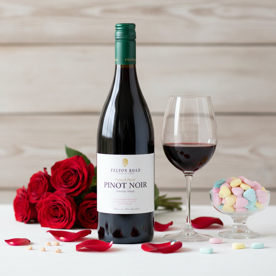 Load image into Gallery viewer, A bottle of Felton Road Cornish Pinot Noir red wine stands on a white surface beside a filled wine glass, red roses, candy in a glass bowl, and scattered red rose petals, against a light wood plank background.