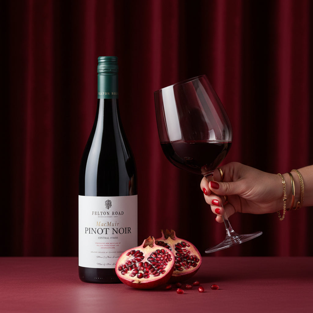 Load image into Gallery viewer, A bottle of Felton Road MacMuir Pinot Noir red wine stands on a dark red surface with sliced pomegranates and a hand holding a filled wine glass against a deep red curtain backdrop.