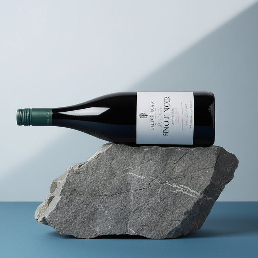 Load image into Gallery viewer, A bottle of Felton Road MacMuir Pinot Noir wine from the French Wine Institute rests horizontally on a rough grey stone against a clean studio background with a blue surface and soft diagonal shadows.