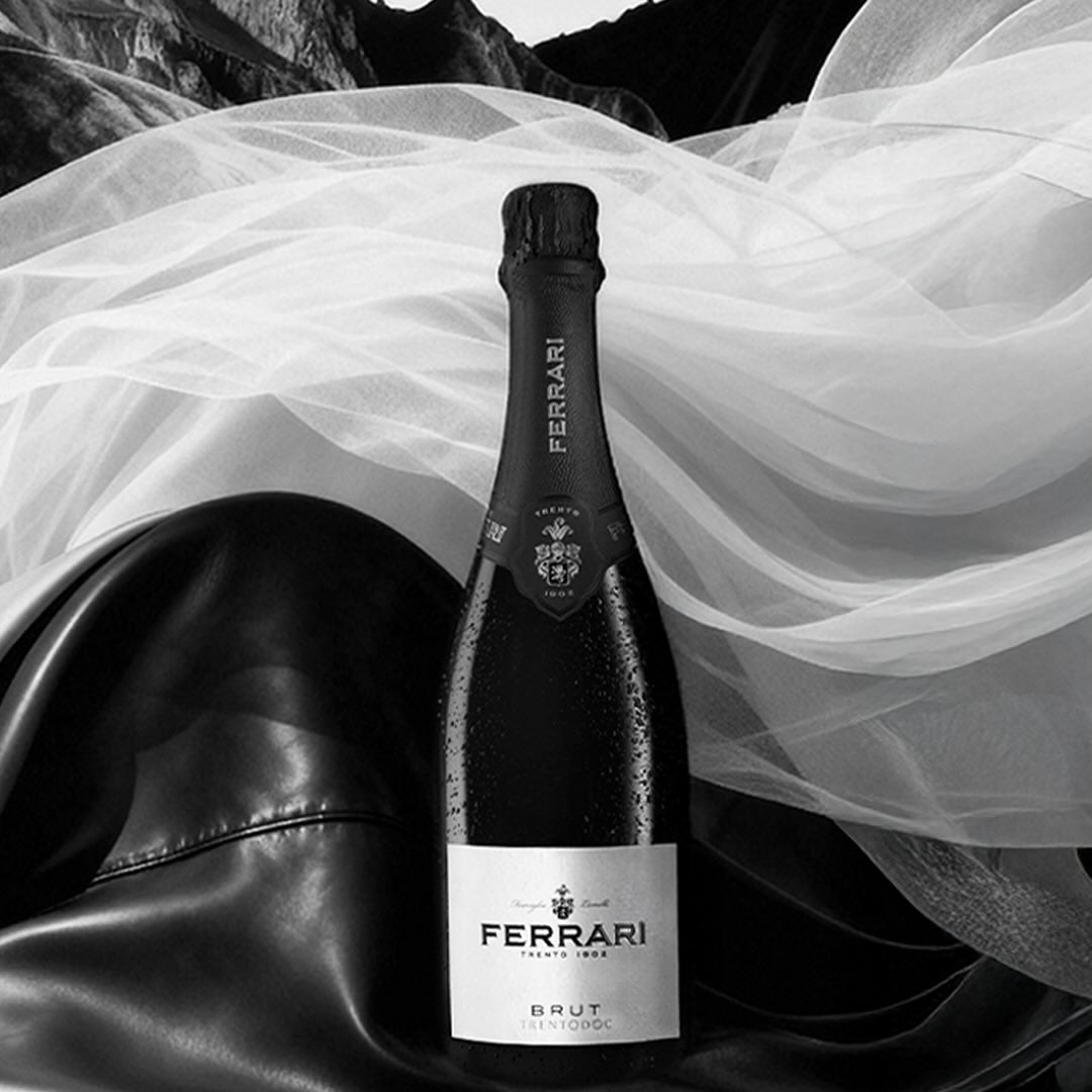Load image into Gallery viewer, Ferrari Trento Brut Metodo Classico sparkling wine bottle on a reflective surface with draped fabric and mountain background