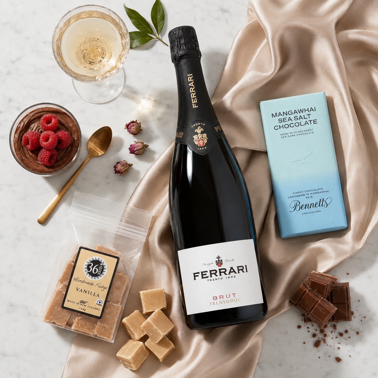 Load image into Gallery viewer, A luxury gift assortment featuring a bottle of Ferrari Trento Brut Metodo Classico in the centre, surrounded by gourmet ingredients and treats on a white marble surface: a packet of 36 Degrees vanilla fudge, a Bennett’s Mangawhai Sea Salt chocolate bar, chocolate pieces and vanilla fudge pieces, styled on a beige satin drape over a white marble background.