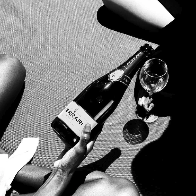 Load image into Gallery viewer, A black and white close-up of a hand holding a bottle of Ferrari Brut sparkling wine next to a wine glass on a textured surface.