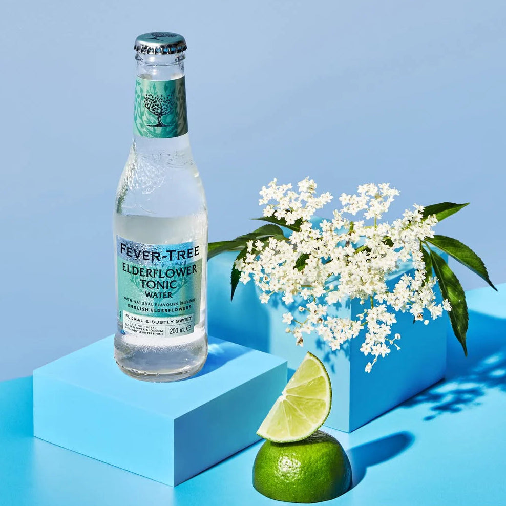 Load image into Gallery viewer, Fever-Tree elderflower tonic water bottle with lime and flowers on a blue geometric platform