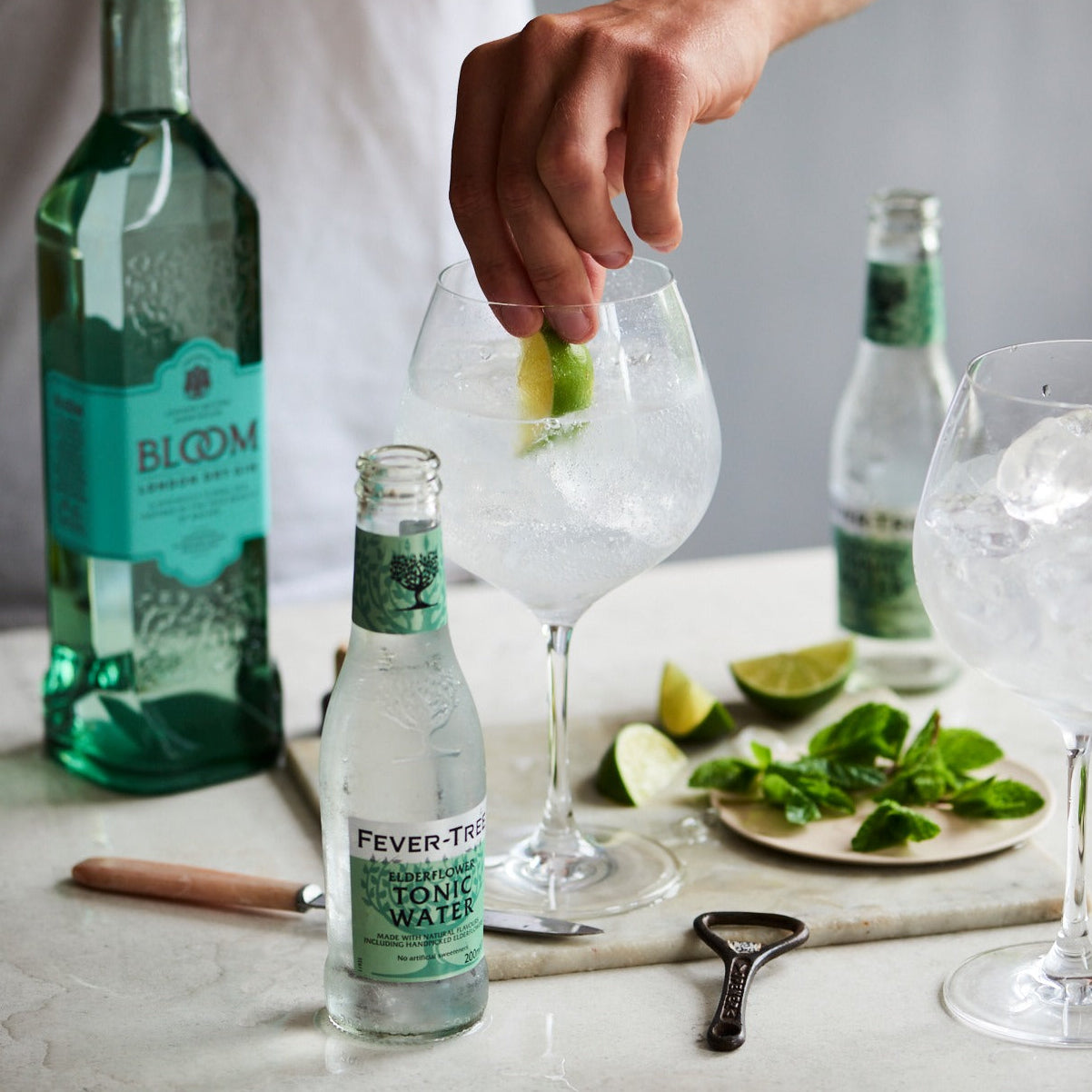 Load image into Gallery viewer, Person preparing a cocktail with bottles of Bloom gin and Fever-Tree tonic water on a table.