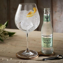 Load image into Gallery viewer, Bottle of Fever-Tree Elderflower tonic water next to a glass with ice and a lemon twist on a wooden surface.