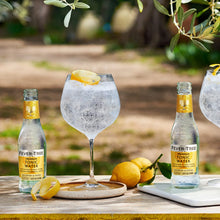 Load image into Gallery viewer, Two bottles of Fever-Tree Indian tonic water with glasses filled with a clear beverage and lemon slices on a wooden table outdoors.