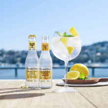 Load image into Gallery viewer, Two bottles of Fever-Tree Indian tonic water and a glass with lemon and mint on a table by a waterfront.