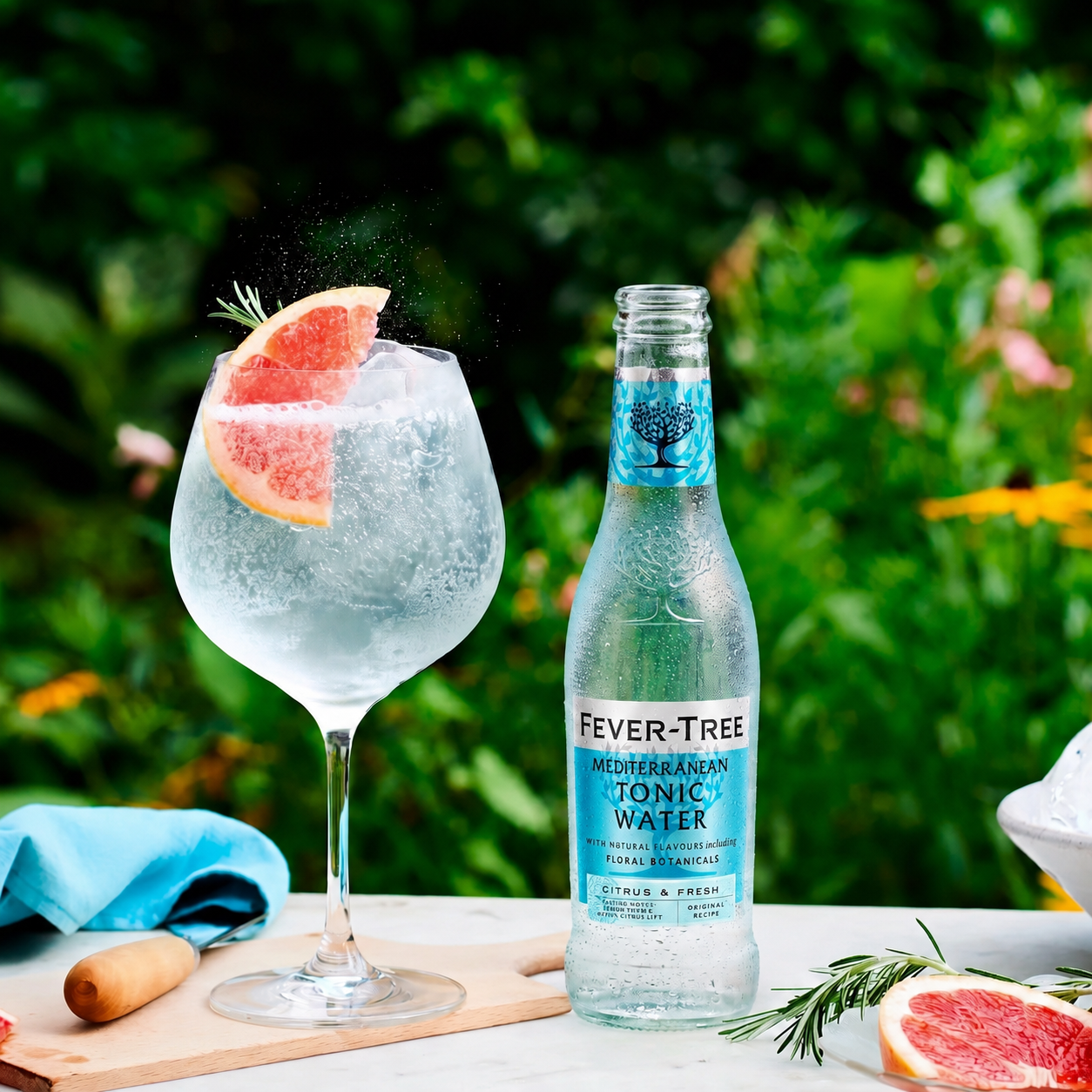 Load image into Gallery viewer, A bottle of Fever-Tree Mediterranean Tonic Water stands next to a large wine glass filled with a clear, carbonated drink and garnished with a grapefruit slice, set on an outdoor table with a blurred green garden background.