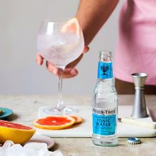 Load image into Gallery viewer, Person holding a glass with ice next to a bottle of Fever-Tree Mediterranean tonic water on a table with fruits.