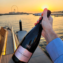 Load image into Gallery viewer, Person opening a bottle of Fleurre de Miraval champagne by the water at sunset.
