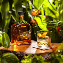 Load image into Gallery viewer, Bottle of Flor de Cana 18 year old Rum on a wooden surface with a cocktail glass in the background, surrounded by greenery.