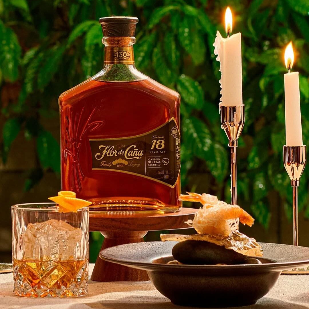 Load image into Gallery viewer, Bottle of Flor de Caña 18-year-old rum with a glass of cocktail and food on a table with candles.