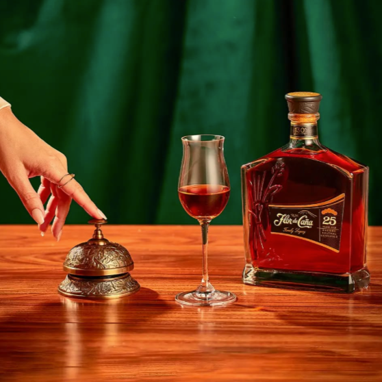 Bottle of Flor de Cana 25 year old Rum with a glass on a wooden table, hand ringing a service bell.