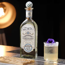 Load image into Gallery viewer, Fortaleza Blanco Tequila bottle with a small glass of cocktail on a wooden coaster, blurred light in the background