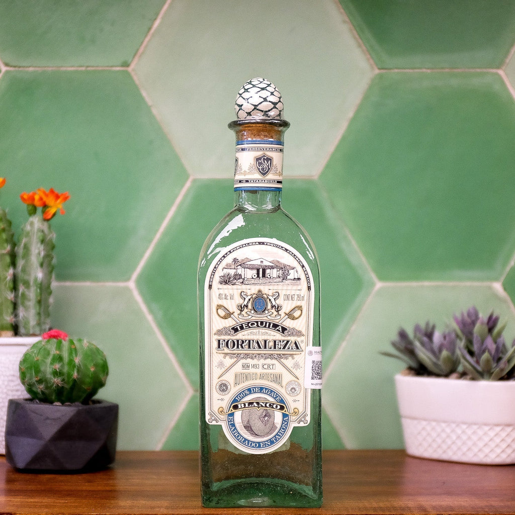 Fortaleza Blanco Tequila bottle with decorative label on a wooden surface with plants and green tiled wall