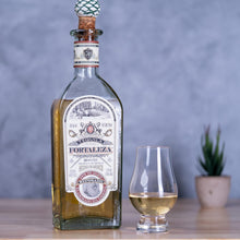 Load image into Gallery viewer, Bottle of Tequila Reposado Fortaleza with a glass of tequila on a wooden surface against a gray background