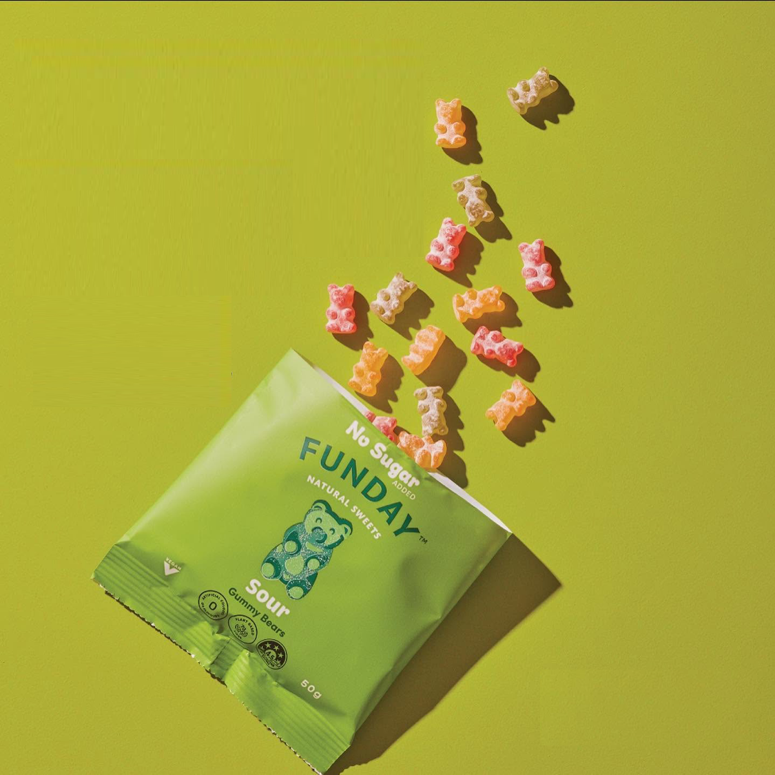 Load image into Gallery viewer, A green bag of Funday Sour Vegan Gummy Bears lies on a bright yellow-green background with several gummies scattered above it in a diagonal arrangement.