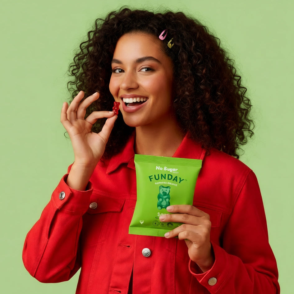 Load image into Gallery viewer, A smiling woman with curly dark hair wears a bright red jacket and holds a green bag of FUNDAY Vegan Sour Gummy Bear while offering a red gummy to the camera against a solid light green background.