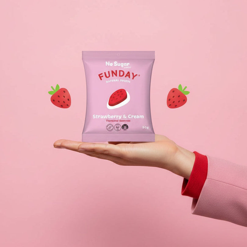 Load image into Gallery viewer, A hand in a pink blazer holds a pink bag of Funday Strawberry &amp; Cream Gummies against a solid pink background, with two small strawberry graphics floating on either side of the bag.
