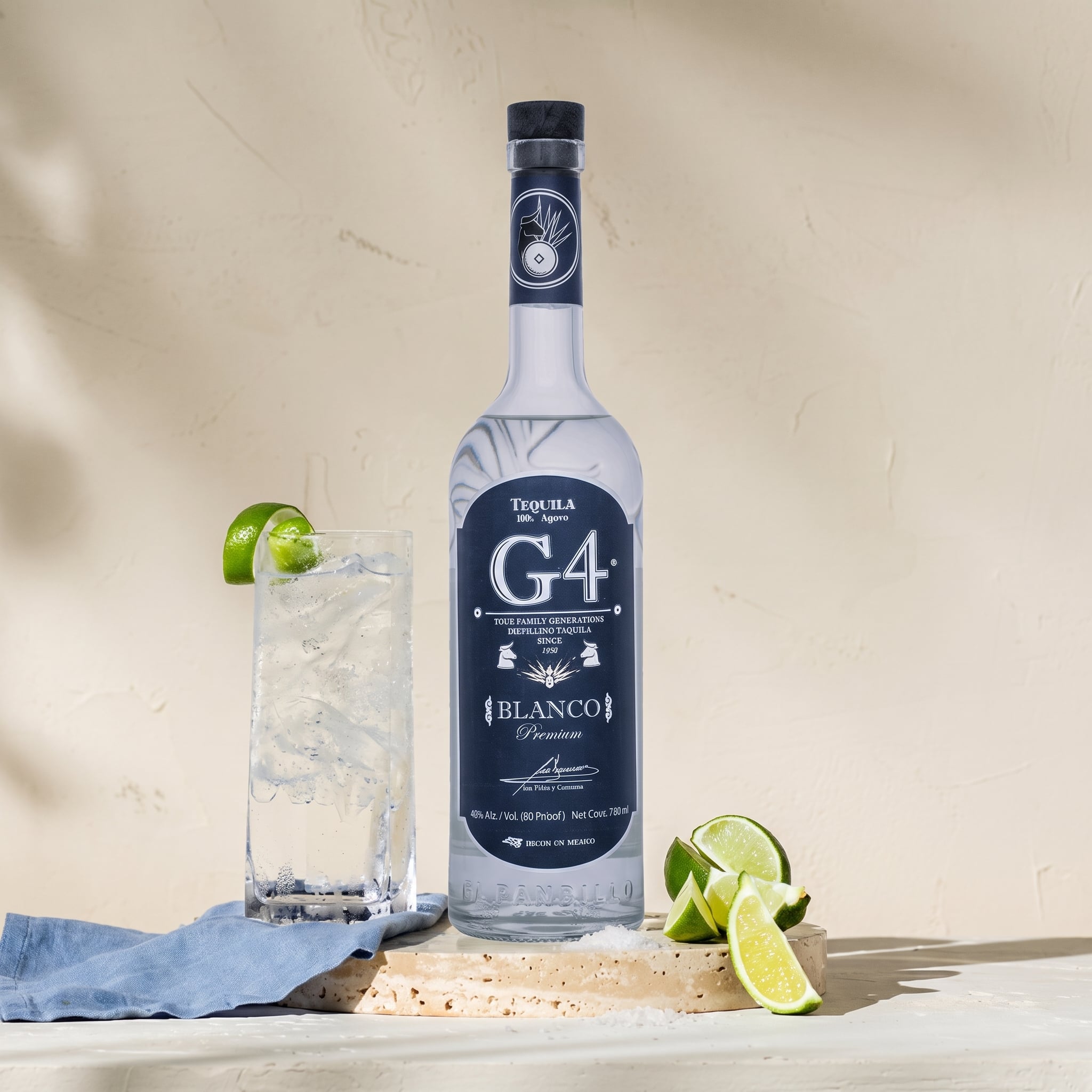 Load image into Gallery viewer, A bottle of G4 Tequila Blanco stands on a wooden board beside a tall glass of iced drink with lime wedges, against a warm beige textured background with soft shadows.