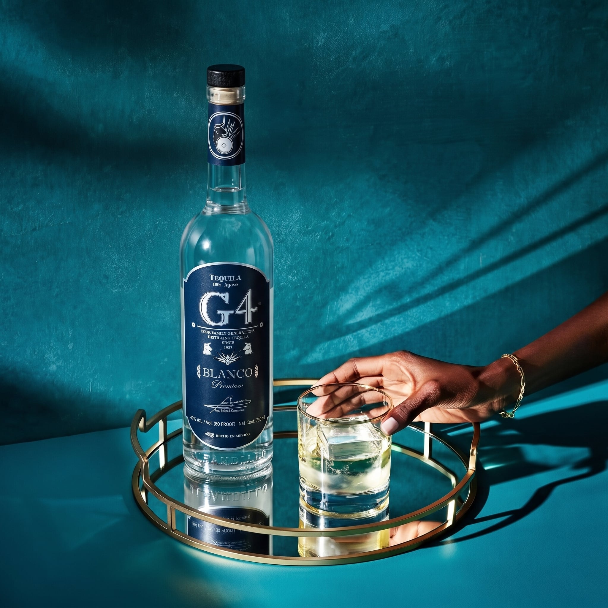Load image into Gallery viewer, A bottle of G4 Tequila Blanco sits on a round gold tray next to a hand holding a glass of the clear liquid, set against a textured teal background.