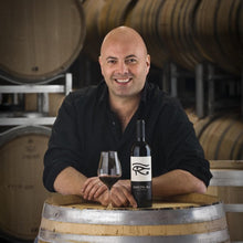Load image into Gallery viewer, Ben Glaetzer sitting on a wine barrel with a Glaetzer-Amon-Ra-Barossa-Shiraz bottle and glass, surrounded by wooden barrels.