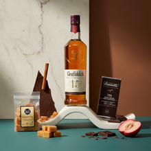 Load image into Gallery viewer, Glenfiddich whisky bottle with Bennetts Dark chocolate, 36 Degrees vanilla fudge and fruit on a decorative stand against a marble wall.