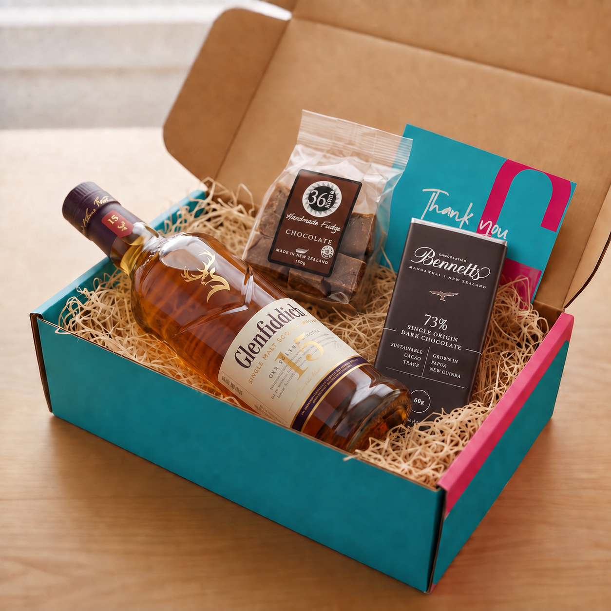 Load image into Gallery viewer, A curated gift box set on a wooden table, featuring a bottle of Glenfiddoch 15 Year Single Malt Scotch, a bag of 36 degrees Chocolate fudge, a box of Bennetts 73% Dark Chocolate, and a teal and pink thank you card inside a turquoise and pink COVE 27 gift box filled with wood-wool.