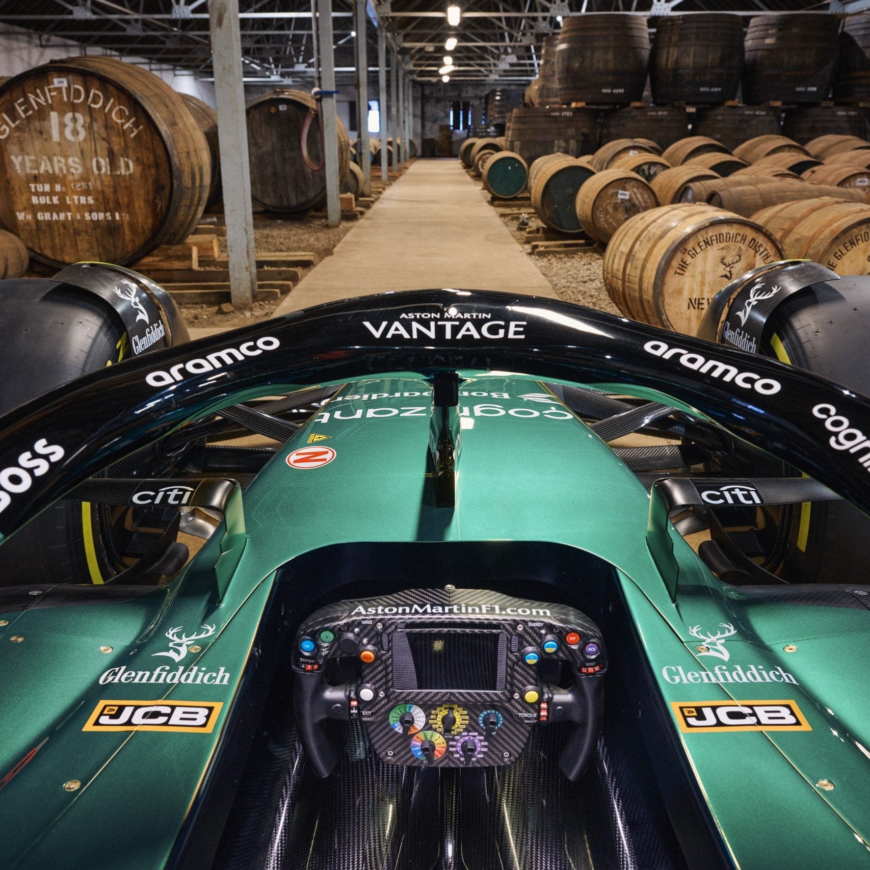Load image into Gallery viewer, An image from the driver's point of view as if sitting in an Aston Martin Formula 1 car with various sponsor logos looking out into theGlenfiddich distillery setting with wooden whisky barrels.