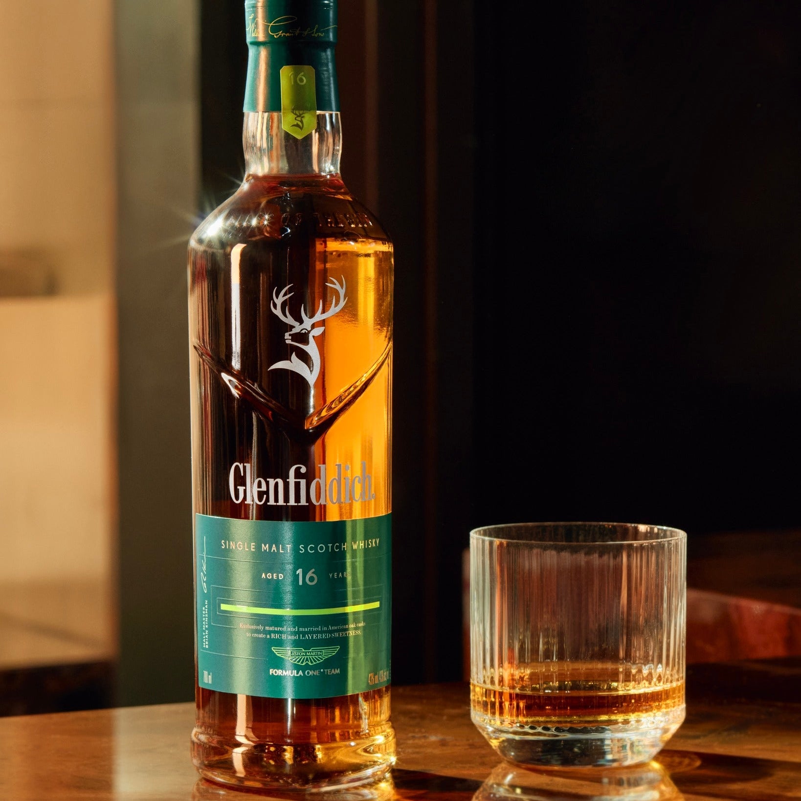 Load image into Gallery viewer, Glenfiddich 16 Year Old Aston Martin F1 Limited Edition bottle with a glass on a wooden surface