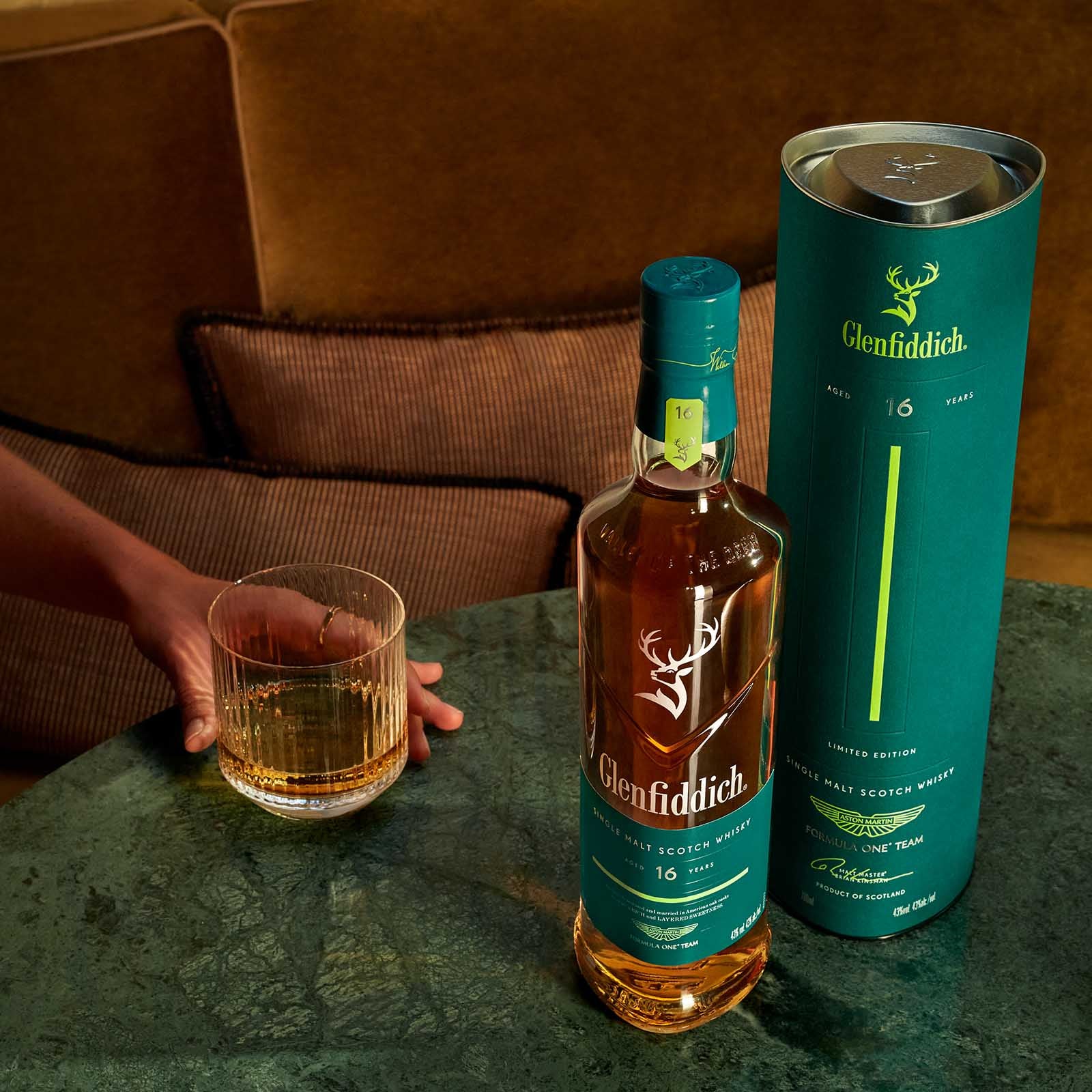 Load image into Gallery viewer, Person holding a glass of neat whisky next to a Glenfiddich 16 Year Old Aston Martin F1 Limited Edition bottle and its glamorous gift box.
