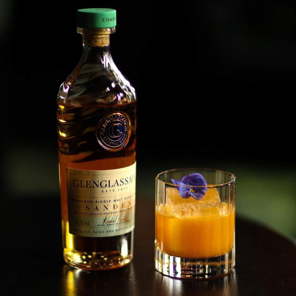 Bottle of Glenglassaugh Sandend whisky and a cocktail with a purple flower on a dark surface.