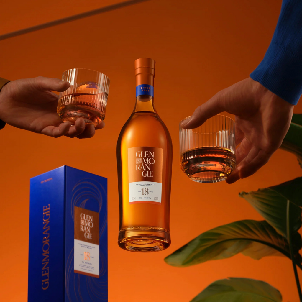 Two hands holding Glenmorangie 18 year old whisky glasses with a bottle and box on an orange background