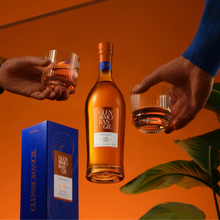 Load image into Gallery viewer, Two hands holding Glenmorangie 18 year old whisky glasses with a bottle and box on an orange background