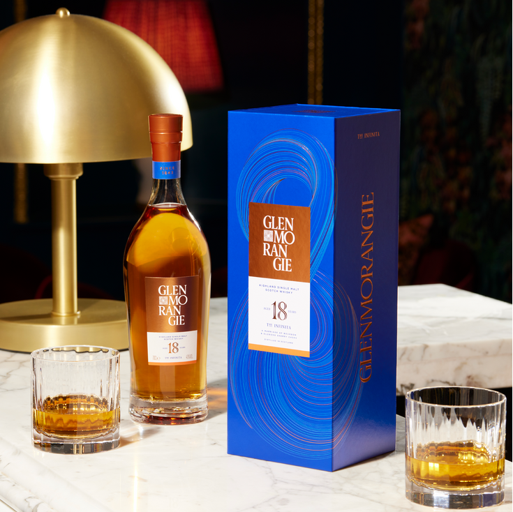 Glenmorangie 18 year old whisky bottle, glass, and packaging on a table with a gold lamp in a dimly lit room.