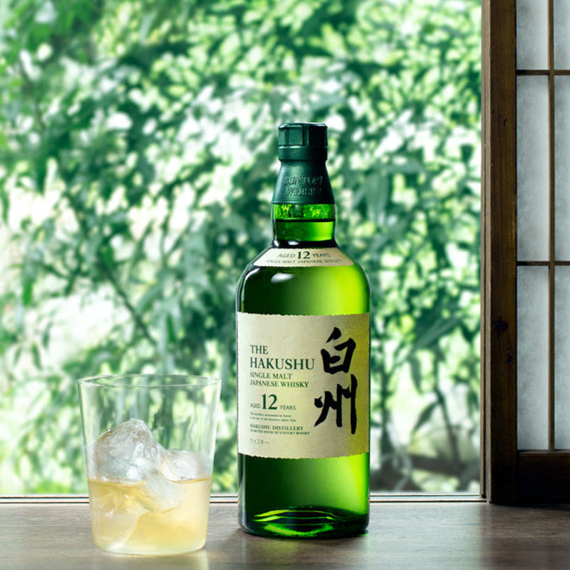 Bottle of Hakushu whisky with a glass of whiskey on a windowsill with greenery outside.