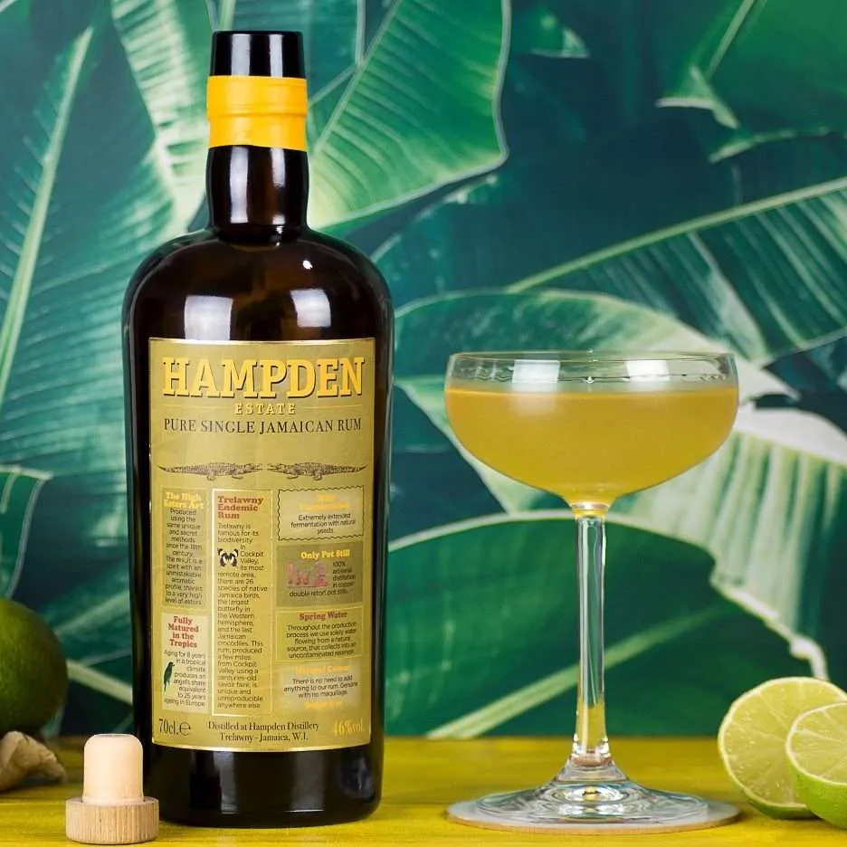 Load image into Gallery viewer, Bottle of Hampden rum with a cocktail glass filled with a yellow drink against a tropical leaf background.