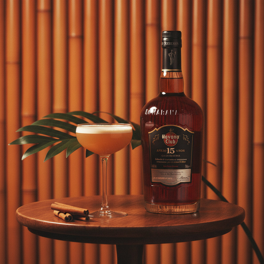 Load image into Gallery viewer, A bottle of Havana Club 15 Year Old Gran Reserva Rum alongside a coupe glass filled with an orange cocktail, set on a wooden table with cinnamon sticks and a green leaf against a warm bamboo background.