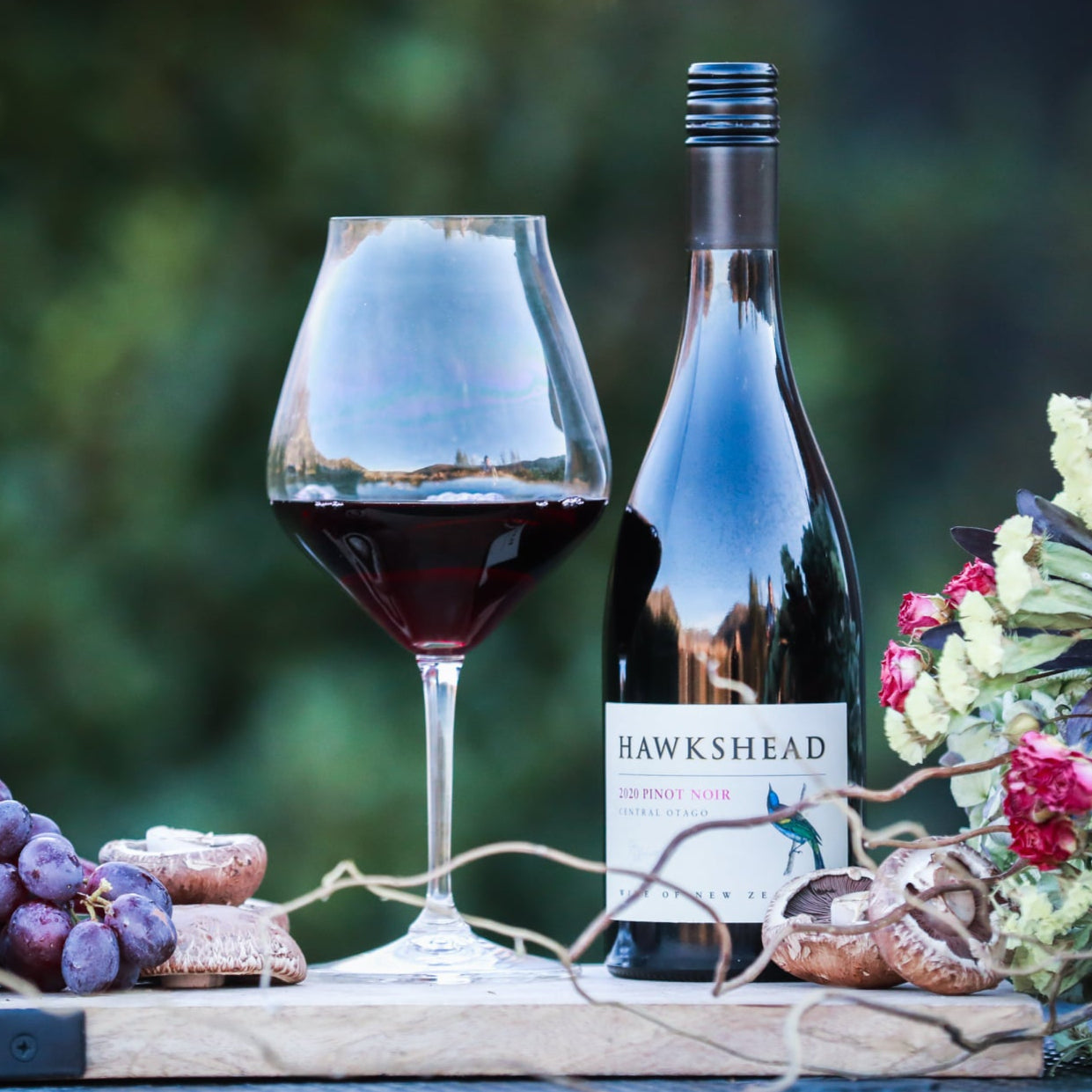 Load image into Gallery viewer, Wine bottle, glass, and grapes on a wooden board with a blurred natural background