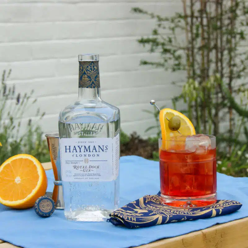 Load image into Gallery viewer, Hayman’s Royal Dock Navy Strength Gin bottle with a cocktail and garnishes on a blue cloth outdoors.