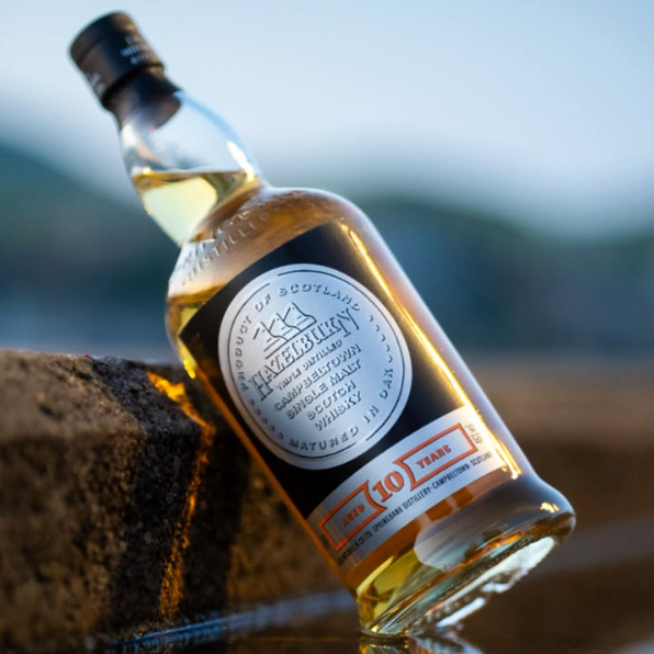 Load image into Gallery viewer, A bottle of Hazelburn 10 year old Single Malt Scotch whisky, 10 Years, leaning against a dark stone surface with a blurred outdoor landscape background.
