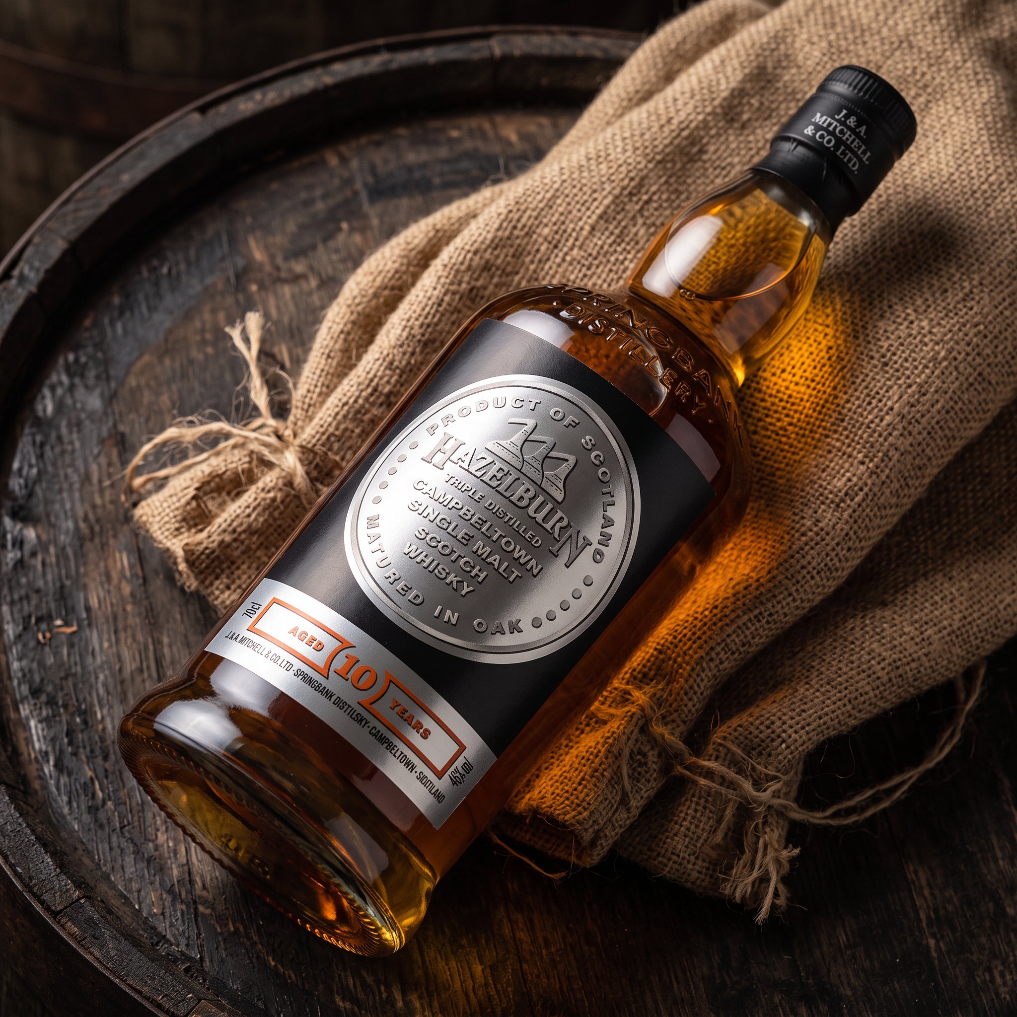 Load image into Gallery viewer, A bottle of Hazeburnn 10 Year Old Single Malt Whisky resting diagonally on a rustic dark wooden barrel, draped over a piece of natural burlap fabric.