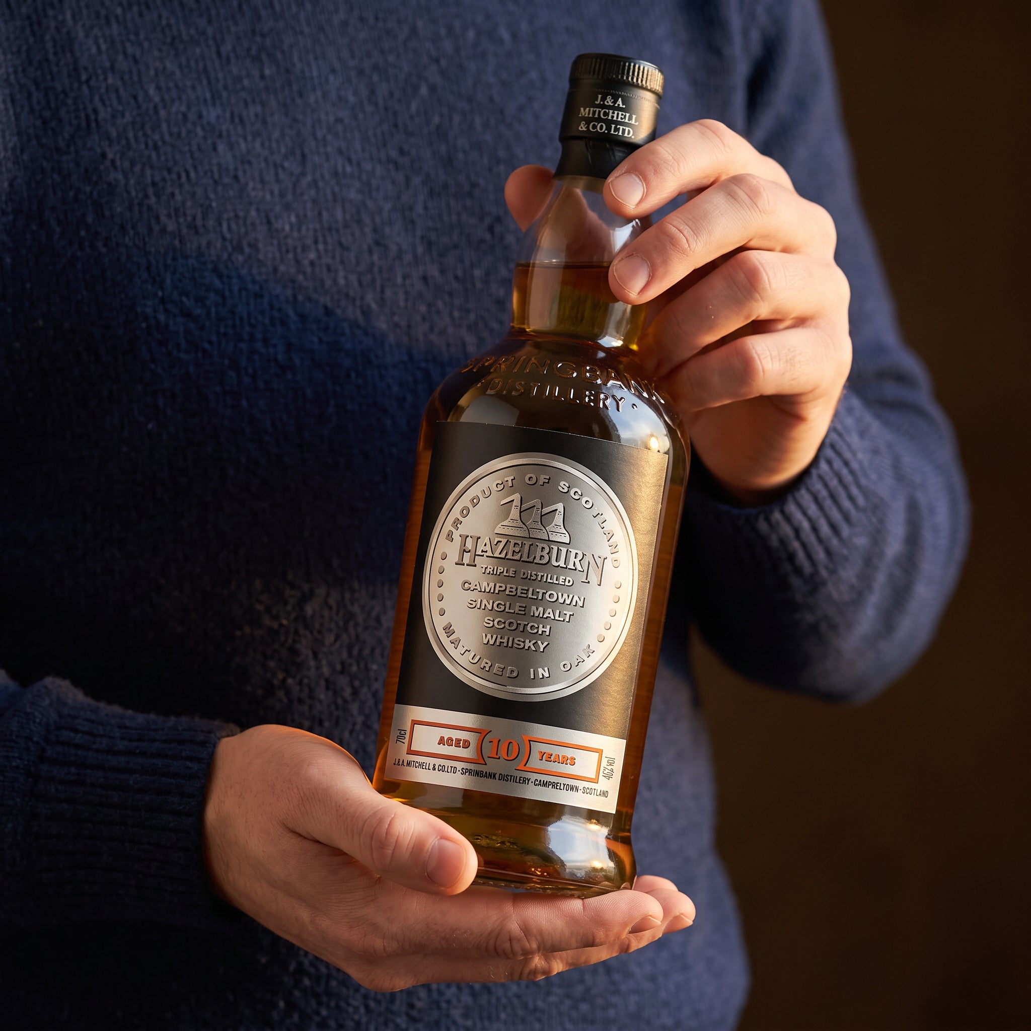 Load image into Gallery viewer, A person in a navy blue sweater holds a bottle of Hazelburn 10 Year Old Single Malt Scotch whisky against a dark brown background.