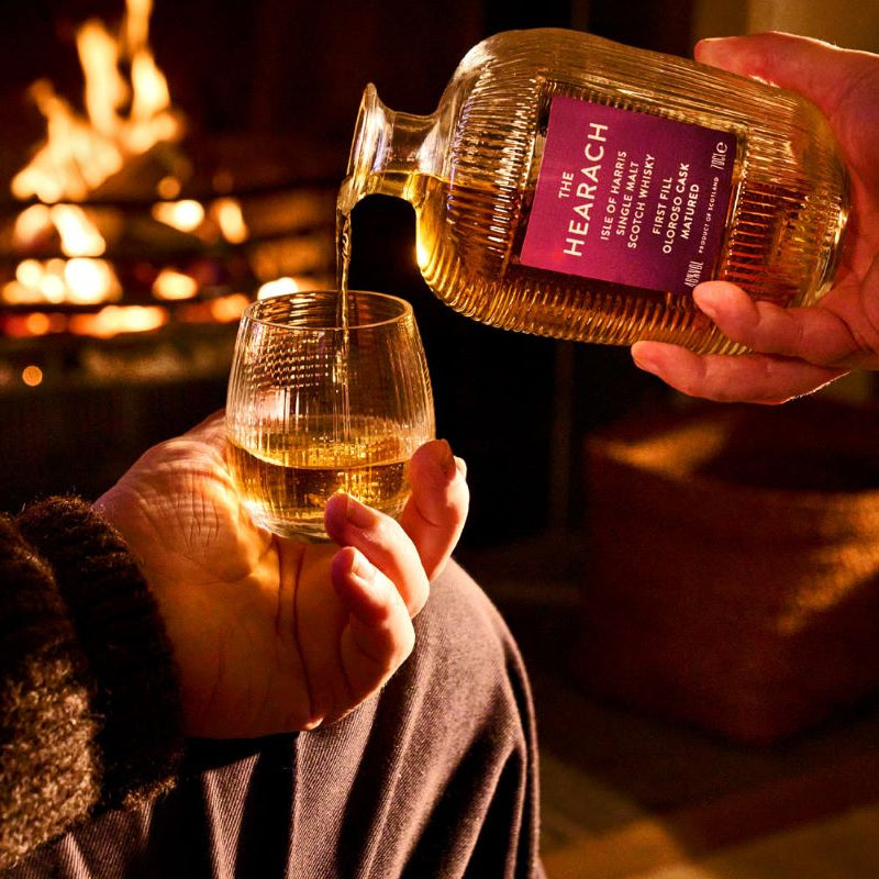 Load image into Gallery viewer, A bottle of The Hearach Oloroso Cask Single Malt Scotch Whisky being poured from a ribbed glass bottle into a matching glass, held by hands in a cozy indoor setting with a warm fireplace in the background.