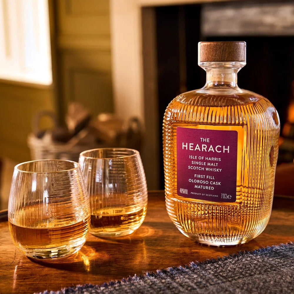 Load image into Gallery viewer, A bottle of The Hearach Oloroso Cask Single Malt Scotch Whisky and two matching stemless glasses filled with whisky, set on a wooden table in a cozy room with a blurred fireplace in the background.