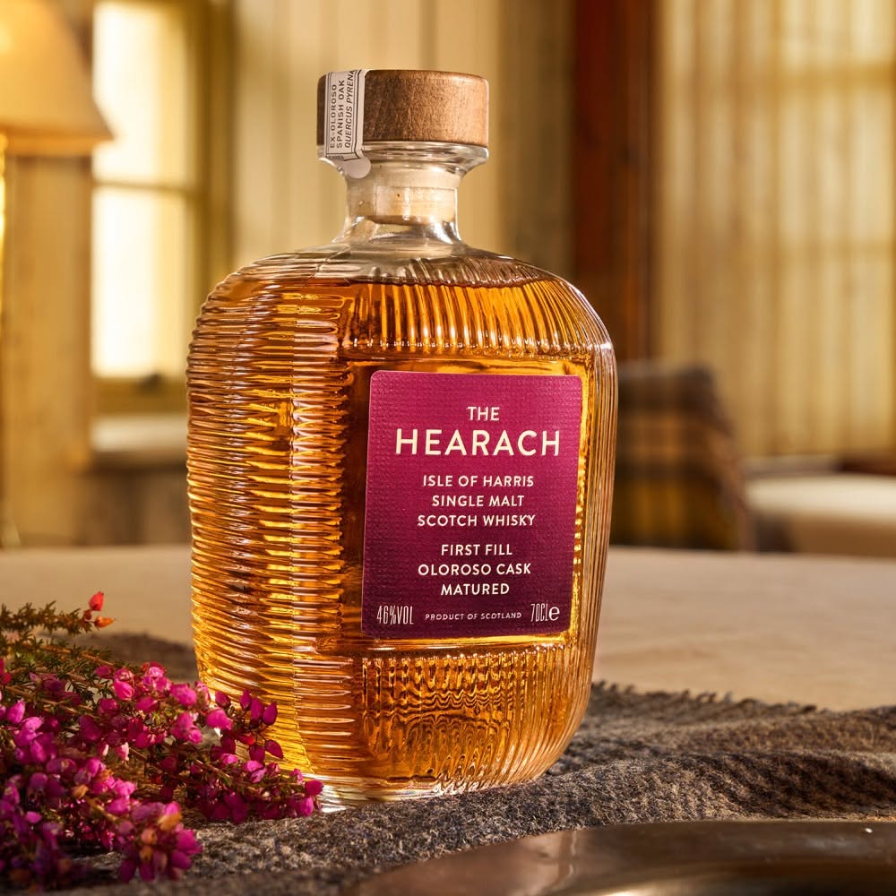 Load image into Gallery viewer, A bottle of The Hearach Oloroso Cask Single Malt Scotch Whisky sits on a rustic table with a textured grey cloth and small pink flowers in the foreground. The background shows a warm, softly blurred interior with wooden barrels and a lamp, creating a cozy, traditional atmosphere.